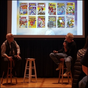 Curator Charles Viera on the left stool next to Patrick McDonnell on the right stool. They're turned around and looking up to see examples of Marvel Comic book covers including Daredevil, X-Men, and Avengers.