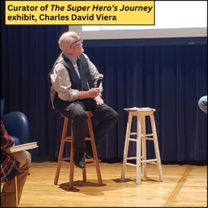 Curator Charles Viera seated on a stool and holding a microphone while he's turned to look up at the screen behind him.